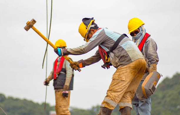 Three construction workers in hard hats and safety gear. The worker in the foreground forcefully swings a large sledgehammer downwards, while another worker carries a welding mask.