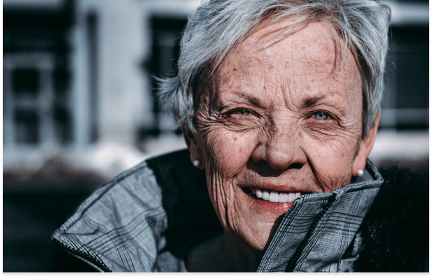 Close-up of a smiling older woman with grey hair, blue eyes, and visible facial wrinkles, wearing a grey jacket and pearl earrings.