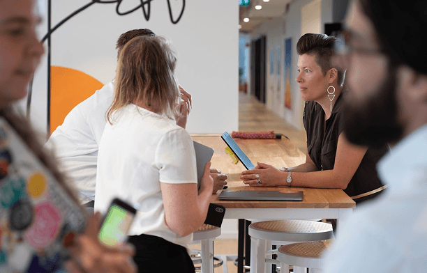 A woman with short hair and distinctive earrings listens intently during a group discussion at a wooden table with laptops in a bright, modern office.