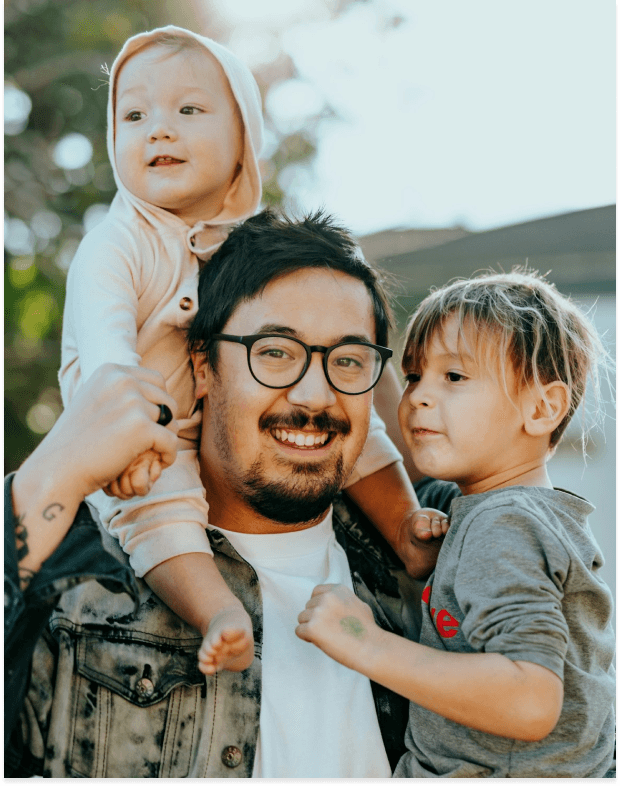 A smiling father in glasses and a denim jacket holds his two young children, one on his shoulders and one in his arm, in a sunny outdoor setting.