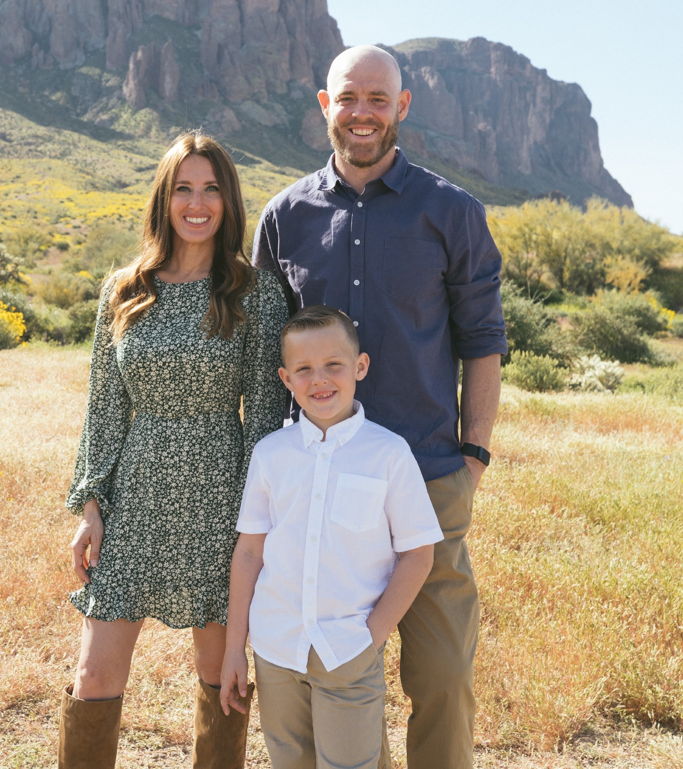 A smiling family of three, a woman, man, and young boy, stand outdoors in a sunny desert landscape with mountains in the background.