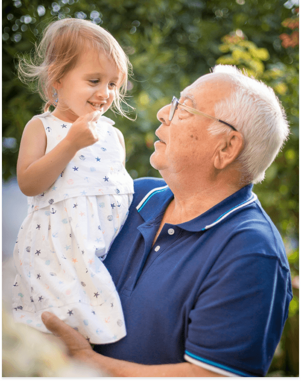 A smiling older man with glasses holds a cheerful young girl in a patterned dress, looking at each other warmly outdoors.
