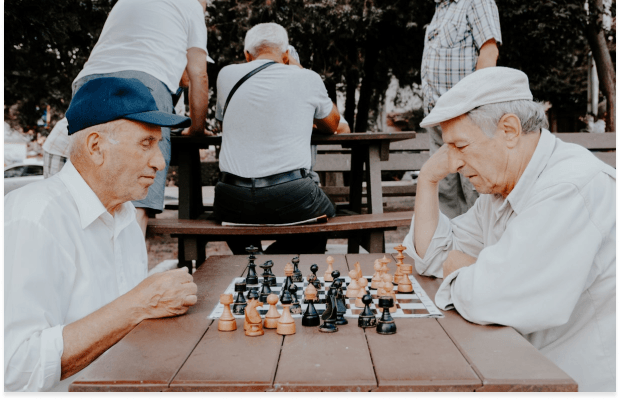 Two older men are engaged in a game of chess outdoors. The man on the left wears a blue cap, while the man on the right, in a light cap, thoughtfully considers his next move.
