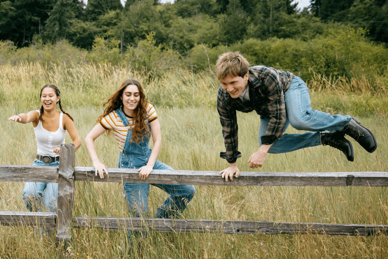 Three smiling young people, two women and a man, actively climbing a rustic wooden fence in a tall grassy field.