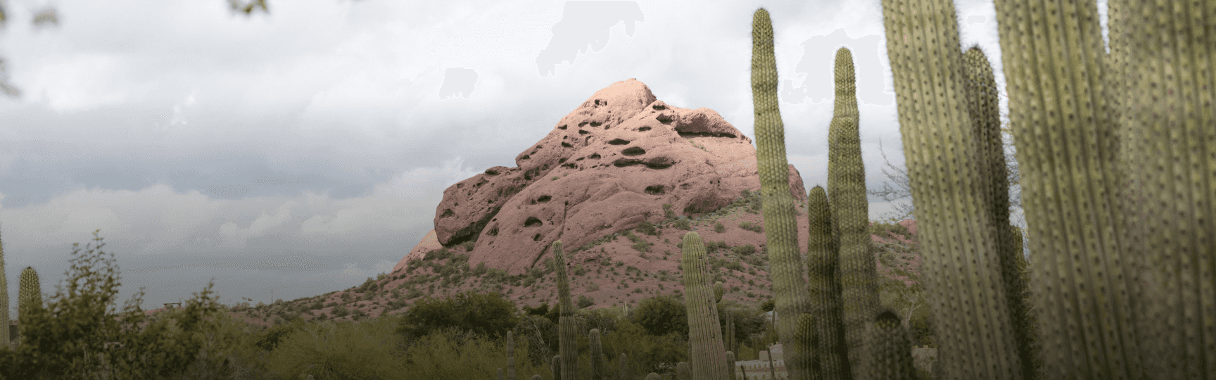 A distinctive red sandstone mountain with numerous eroded holes, flanked by tall saguaro cacti, under a cloudy desert sky.