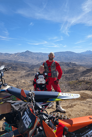 Smiling father and son in full motocross gear pose with their dirt bikes at a scenic desert mountain viewpoint.