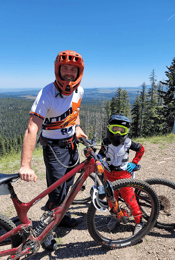 Two mountain bikers, an adult and a young child, stand with their bikes on a sunny mountain summit overlooking a vast forest.