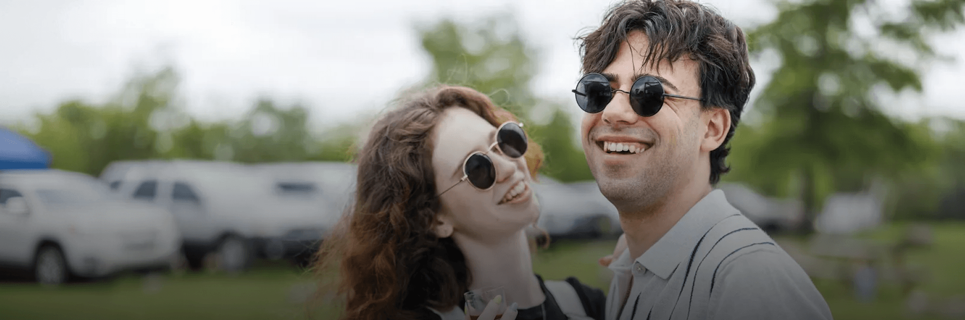 A smiling man in round sunglasses looks forward, while a woman with curly hair and similar sunglasses looks happily up at him at an outdoor event.