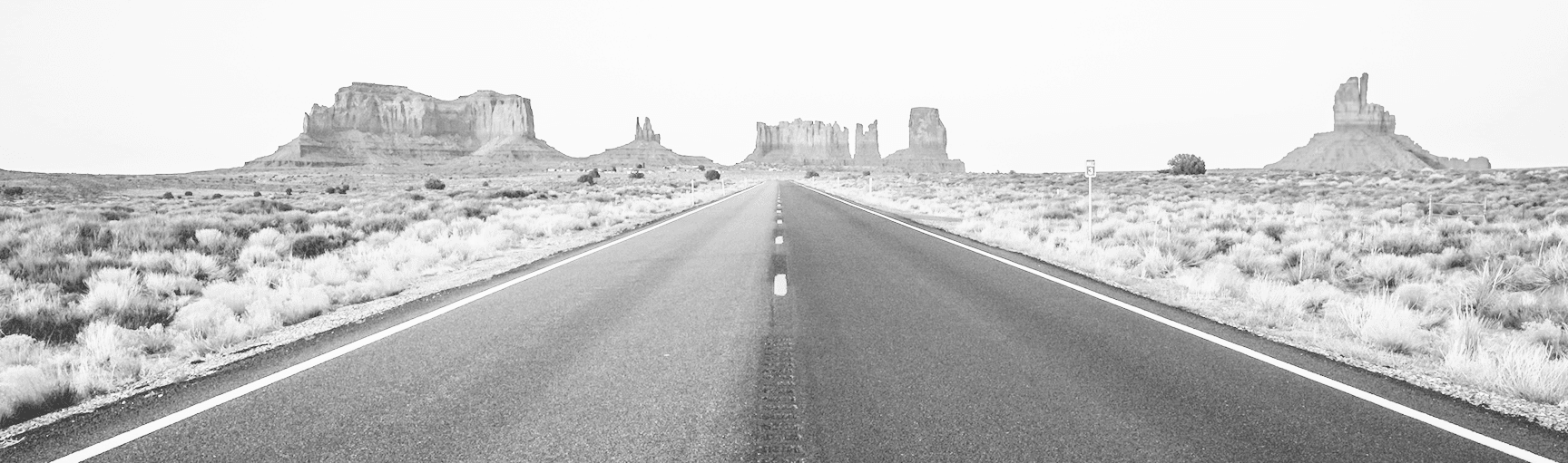Black and white panoramic view of a straight desert road leading towards iconic mesa rock formations in the distance, surrounded by sparse vegetation.