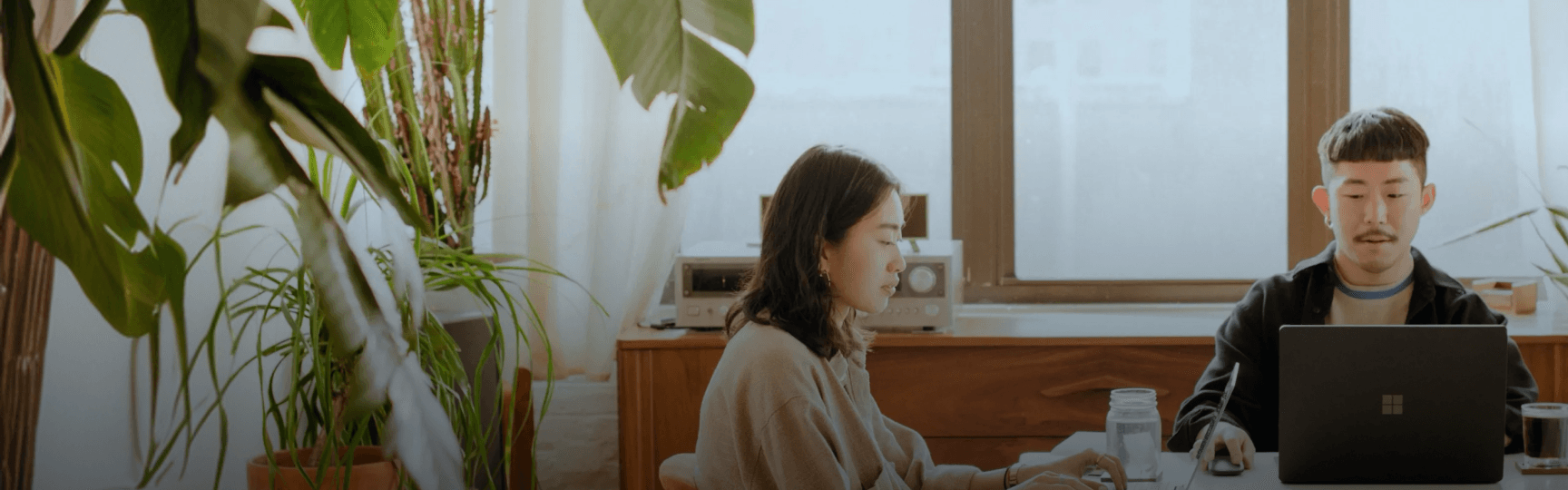Two East Asian individuals, a woman and a man, focused on working on laptops at a wooden desk in a bright, plant-filled room.