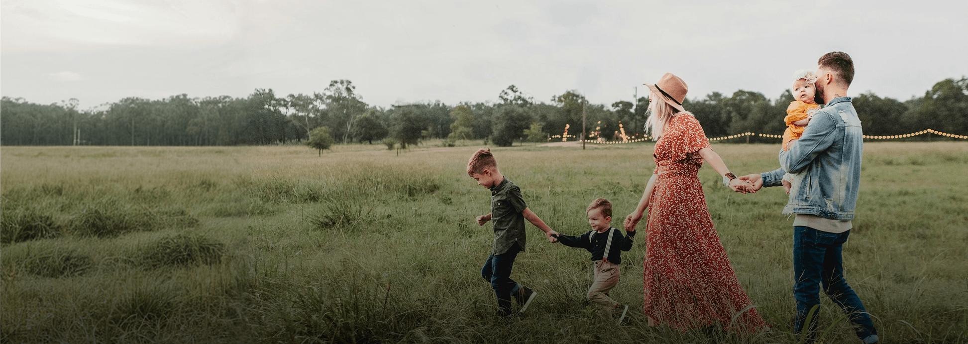 A family of five – a mother, father, two boys, and a baby girl – walks hand-in-hand through a grassy field with trees and string lights in the distance.